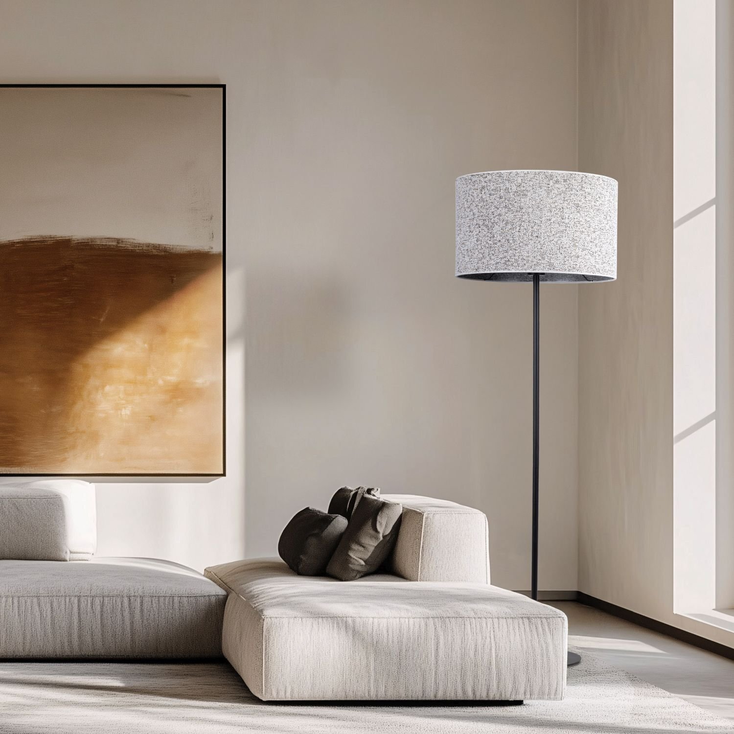 W jasnym wnętrzu znajduje się lampa podłogowa z czarną podstawą i jasnym abażurem. Obok lampy stoi nowoczesna, jasna sofa z ciemnymi poduszkami, a na ścianie wisi duży obraz w odcieniach brązu.