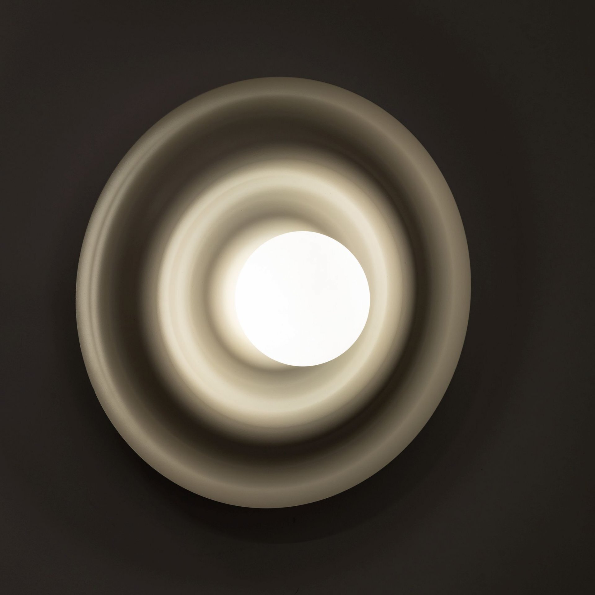 Na obrazie widoczna jest lampa ścienno-sufitowa o okrągłym kształcie z centralnym źródłem światła. Lampa ma faliste, koncentryczne pierścienie w odcieniach szarości.