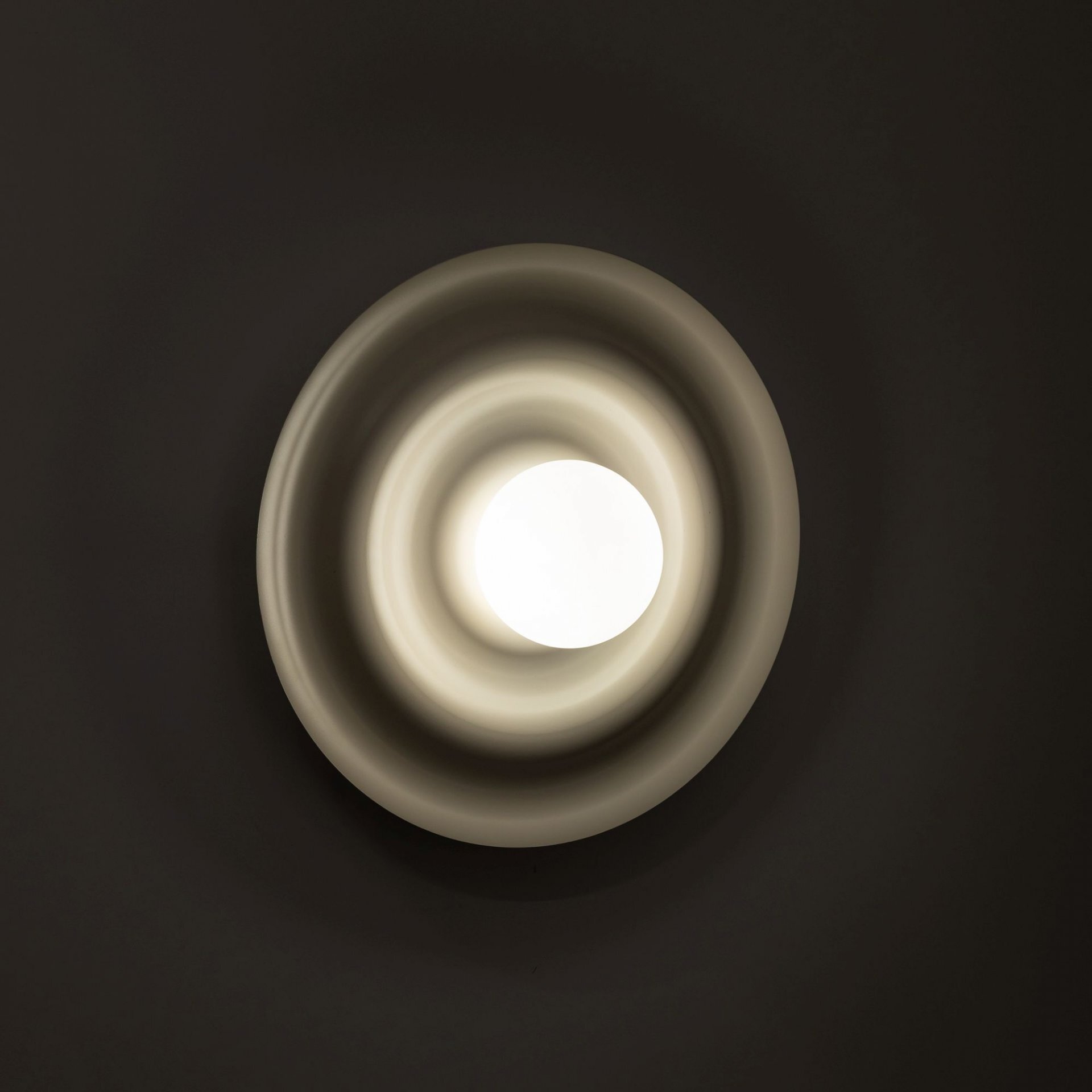 Na ciemnym tle widoczna jest lampa ścienno-sufitowa o okrągłym kształcie z centralnym źródłem światła. Lampa ma falisty wzór wokół źródła światła, co tworzy efekt świetlny na ścianie.