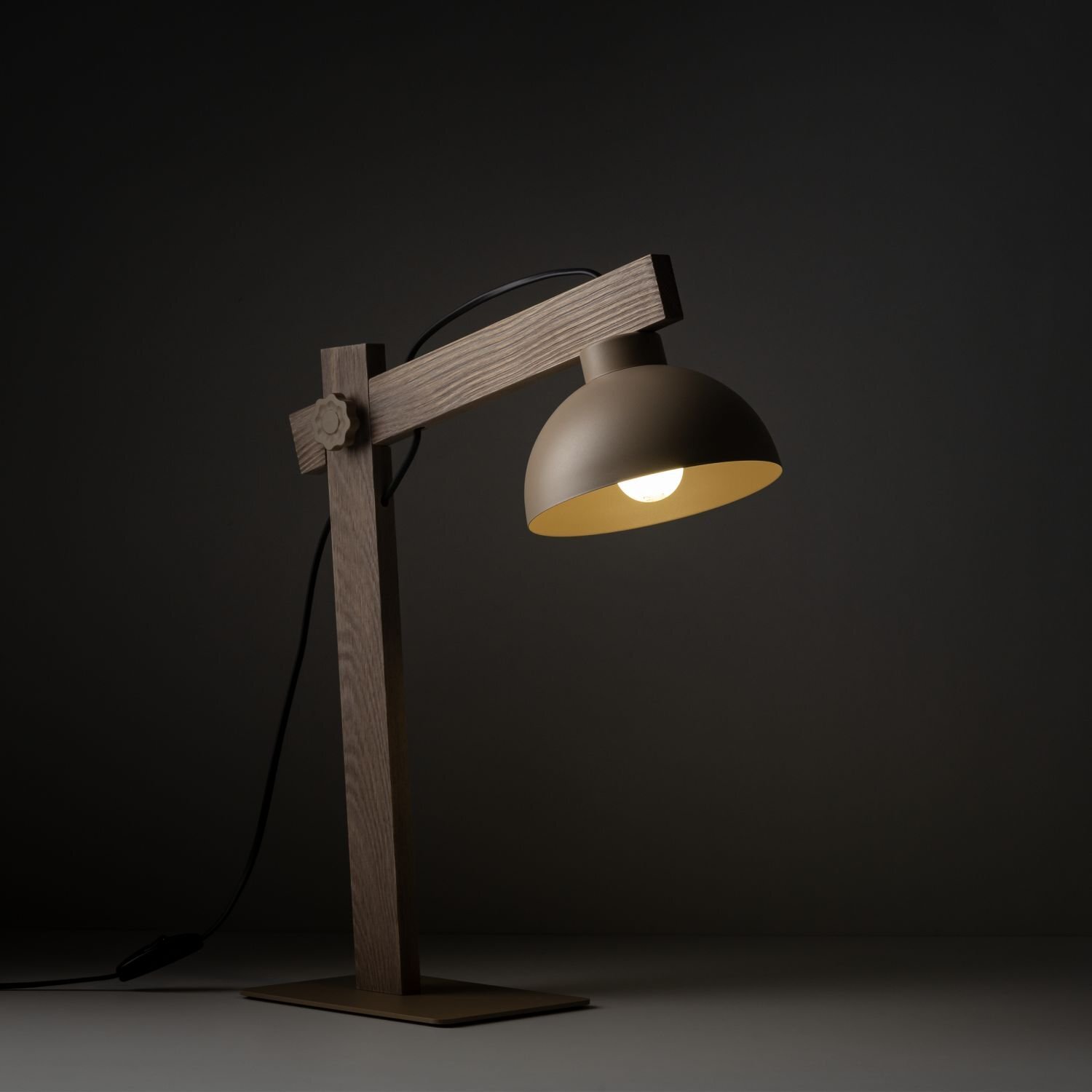 Na obrazie widoczna jest beżowa lampa biurkowa z metalowym kloszem i drewnianą podstawą. Lampa jest włączona, emitując ciepłe światło.