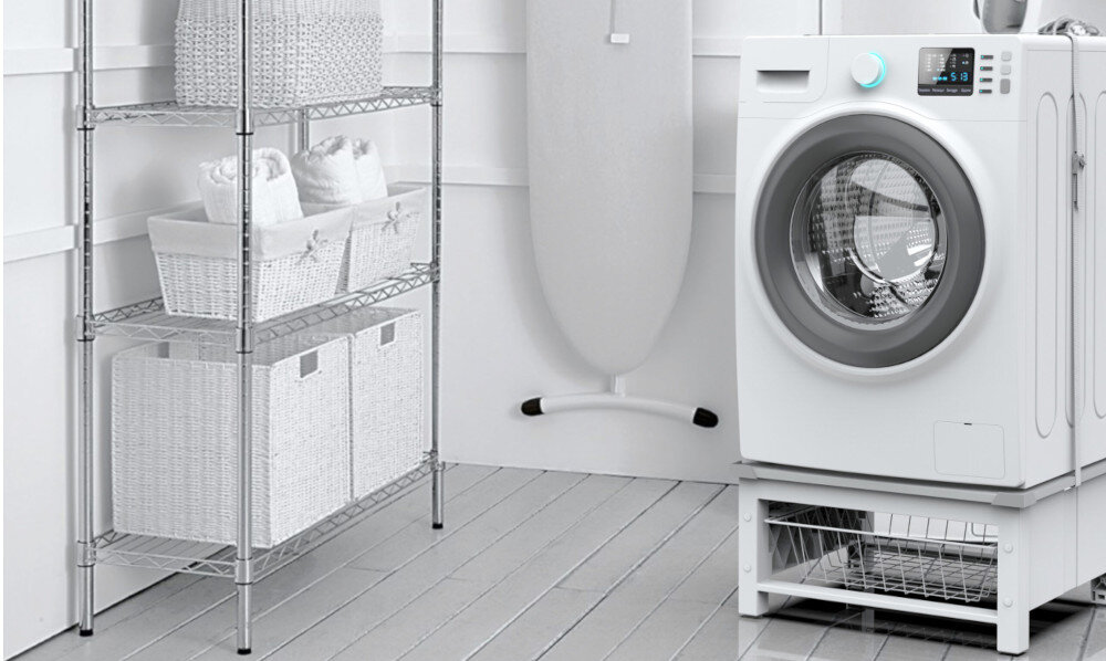 Pralka stoi na podstawie z koszem, obok znajduje się deska do prasowania oraz metalowy regał z wiklinowymi koszami.