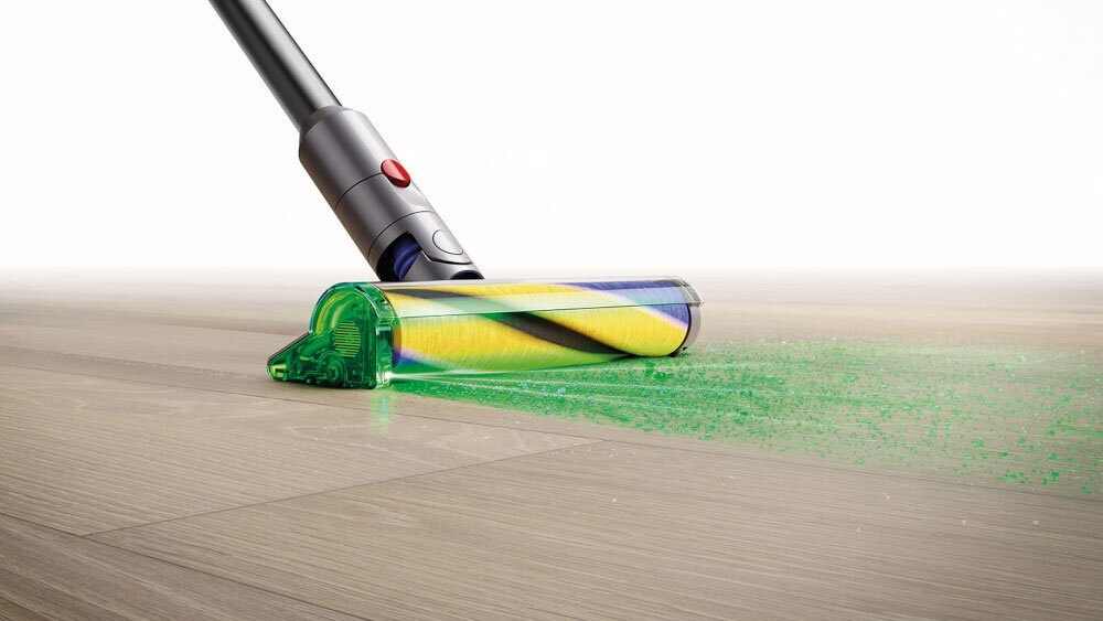 Odkurzacz z przezroczystą głowicą czyszczącą zbiera zielony pył z drewnianej podłogi. W tle widoczne jest jasne, jednolite tło.