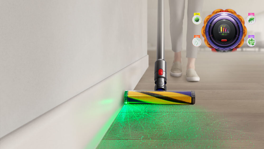 Odkurzacz z zielonym światłem na podłodze, obok ściany. W tle widoczne są nogi osoby oraz okrągły wyświetlacz z kolorowymi ikonami.