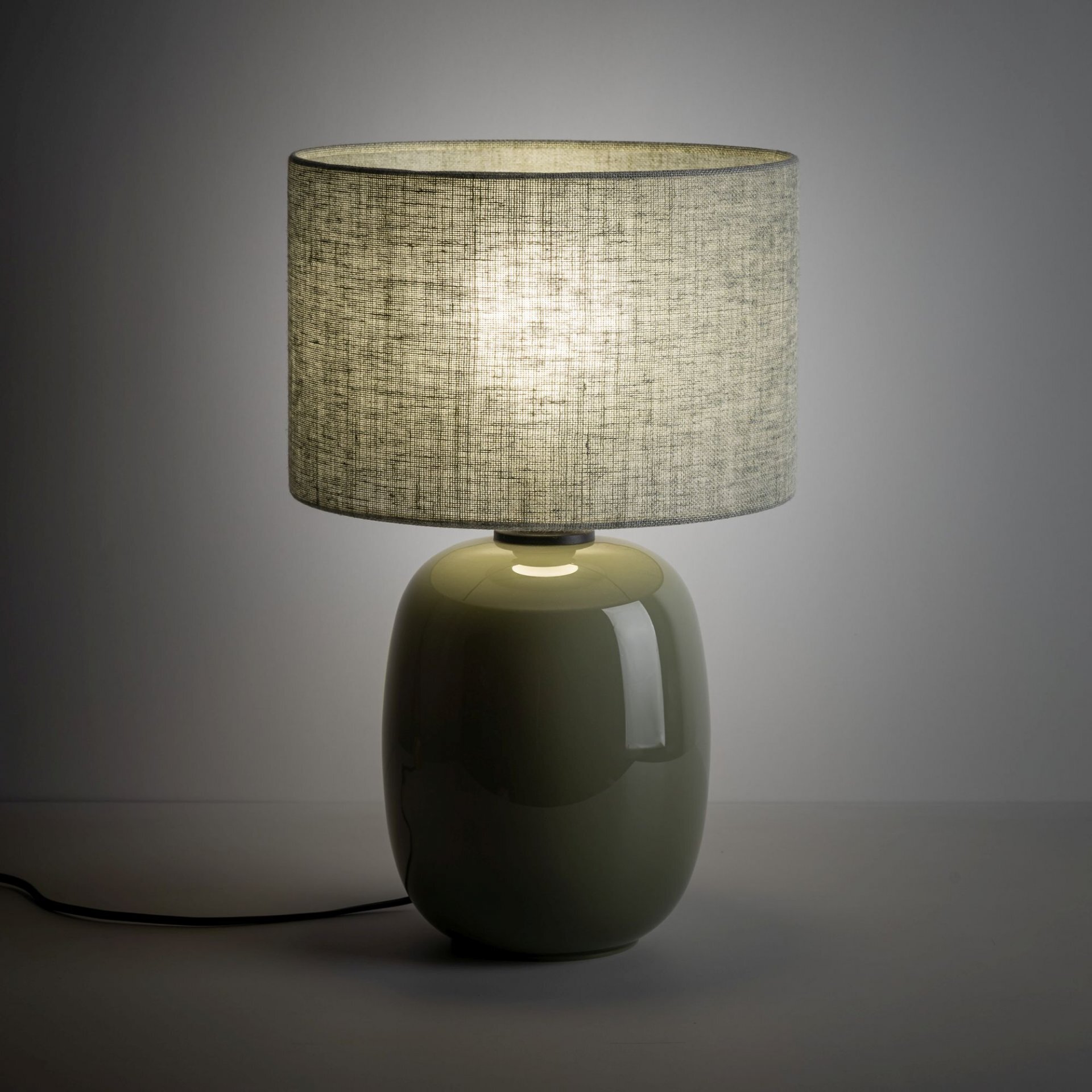 Lampa stołowa z oliwkową, błyszczącą podstawą i lnianym kloszem w szarym odcieniu. Lampa jest włączona, co powoduje delikatne rozproszenie światła przez klosz.