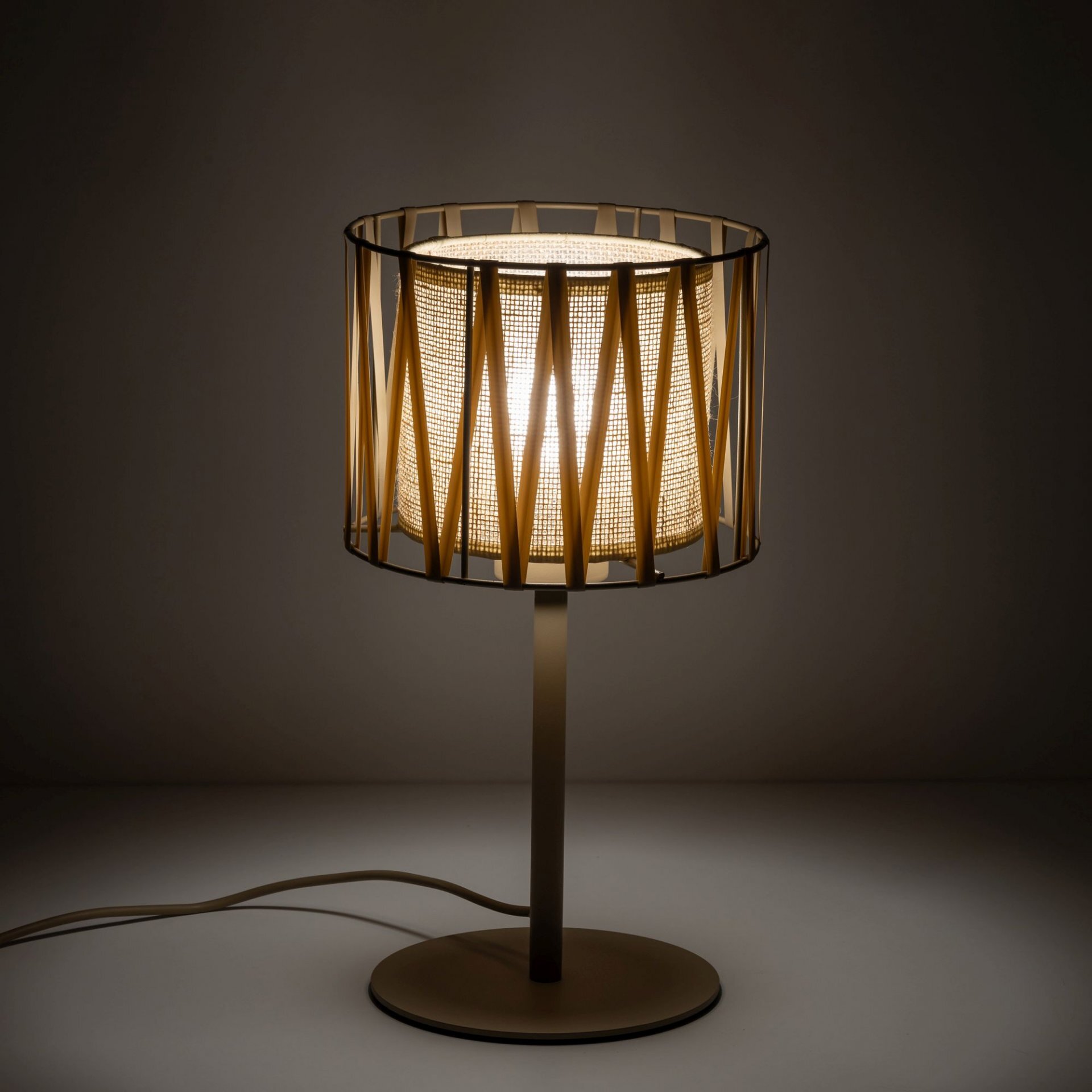 Na obrazie widoczna jest lampa stołowa z beżowym abażurem, który ma ażurową konstrukcję. Lampa stoi na okrągłej podstawie, a jej światło jest włączone, tworząc ciepłą poświatę.