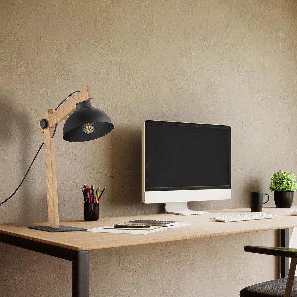 Na biurku znajduje się czarna lampa biurkowa z drewnianym ramieniem oraz komputer z dużym monitorem. Obok monitora stoi kubek, doniczka z rośliną i pojemnik z kolorowymi ołówkami.