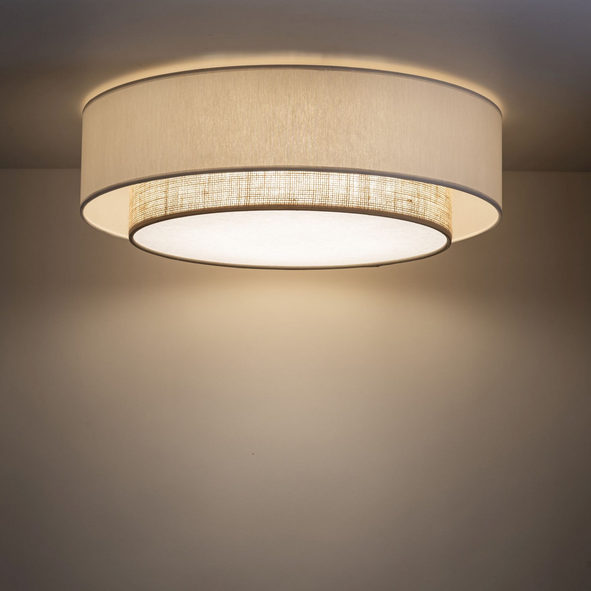Na obrazie widoczna jest lampa sufitowa o cylindrycznym kształcie, z zewnętrzną warstwą w kolorze białym i wewnętrzną z lnianym wzorem. Lampa emituje ciepłe, rozproszone światło.