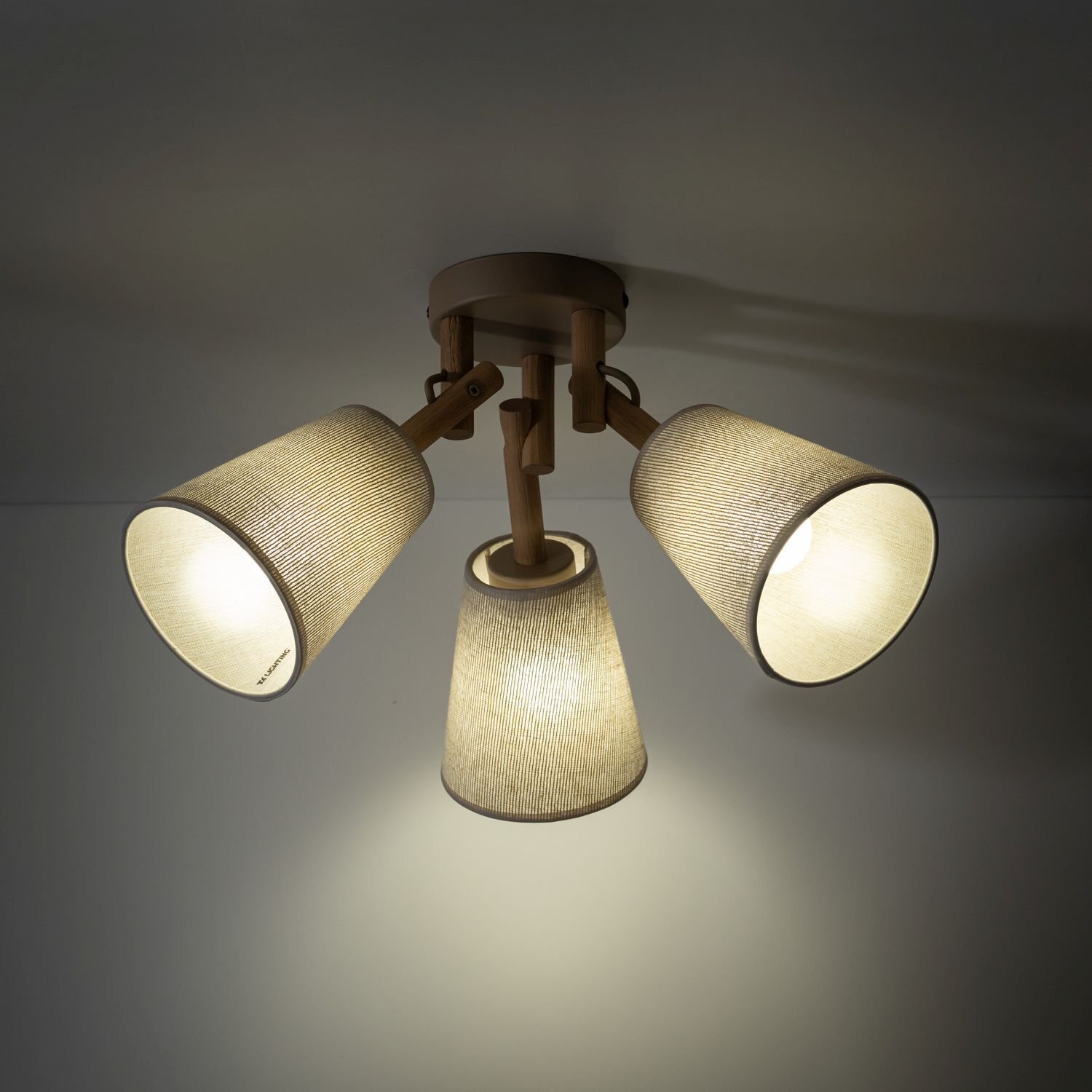 Lampa sufitowa z trzema kloszami w kolorze beżowym, wykonana z materiału przypominającego len, zamontowana na drewnianej podstawie. Klosze są skierowane w różnych kierunkach, a światło jest włączone.