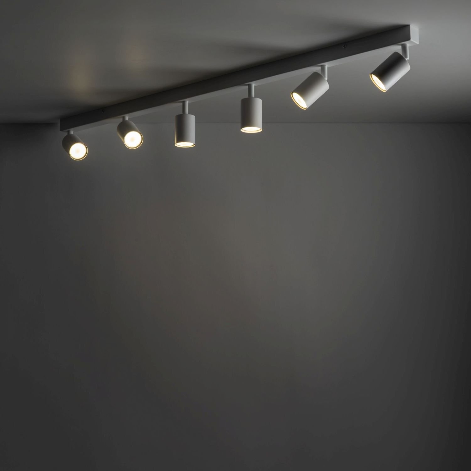 Na obrazie widoczna jest biała lampa sufitowa z sześcioma regulowanymi reflektorami zamontowana na ciemnym suficie. Reflektory emitują ciepłe światło, tworząc nowoczesny wygląd.