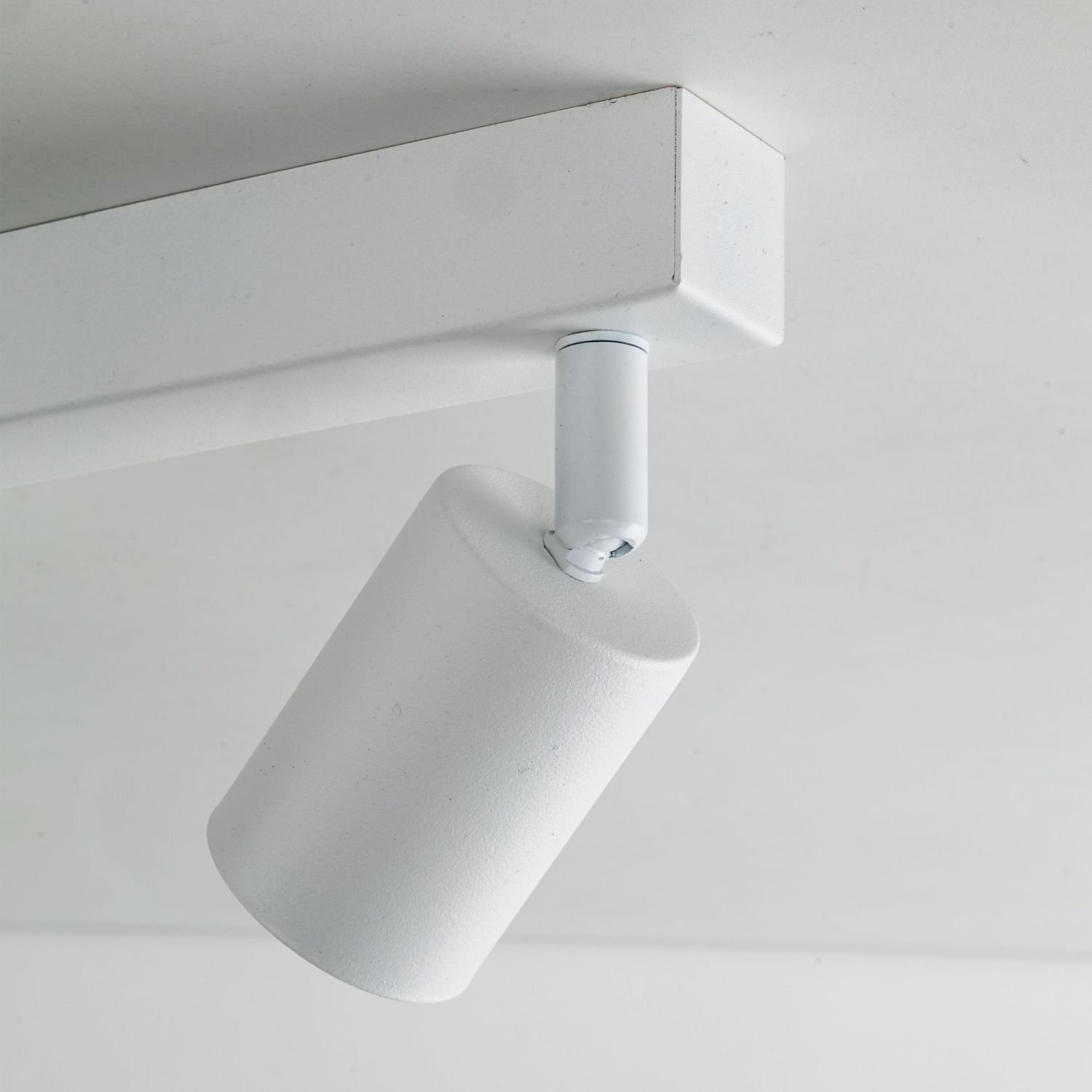 Biała lampa sufitowa z regulowanym reflektorem zamontowana na prostokątnej podstawie. Wykonana ze stali, ma nowoczesny design.