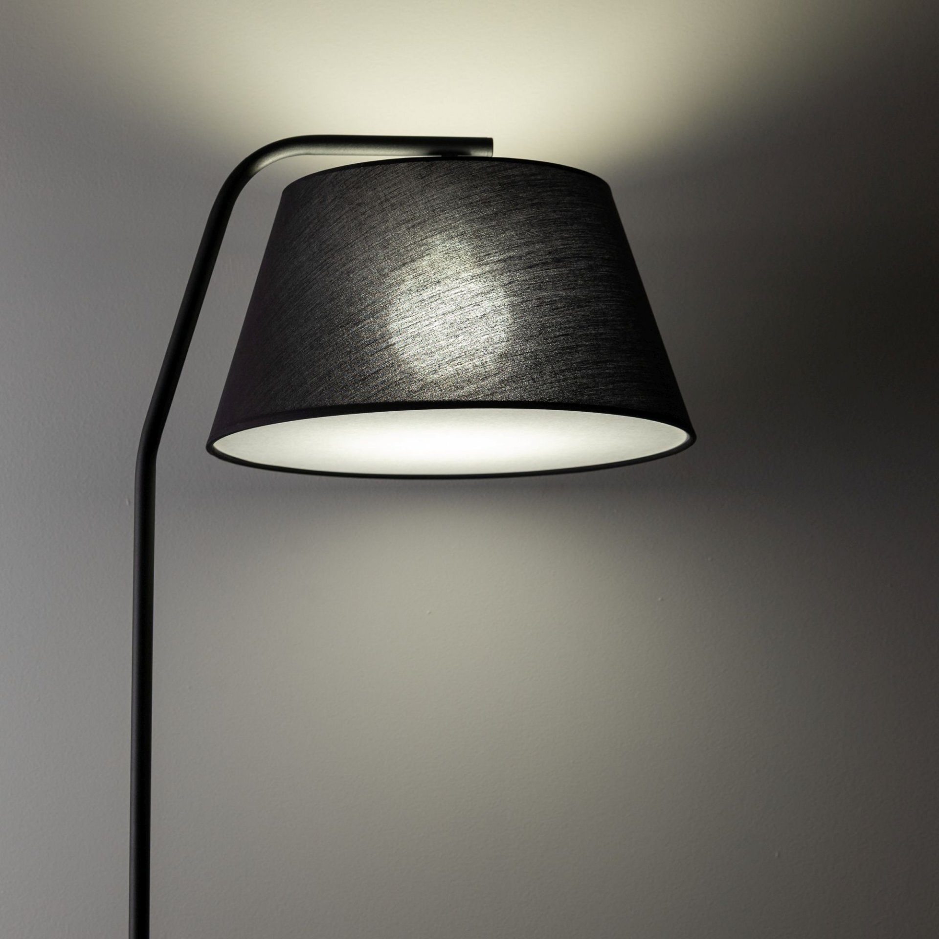 Czarna lampa podłogowa z kloszem w kształcie stożka, emitująca światło na ścianę. Konstrukcja lampy jest nowoczesna i minimalistyczna.