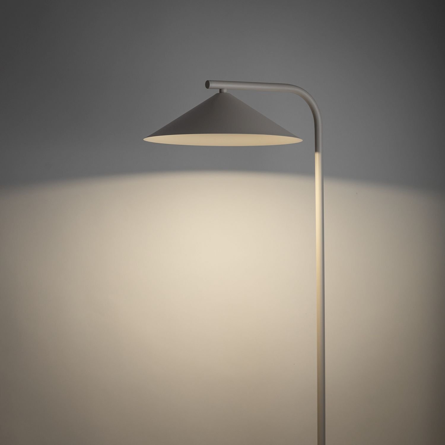Lampa podłogowa z beżowym kloszem w kształcie stożka, emitująca ciepłe światło na ścianę. Konstrukcja lampy jest prosta i nowoczesna, z metalowym ramieniem.