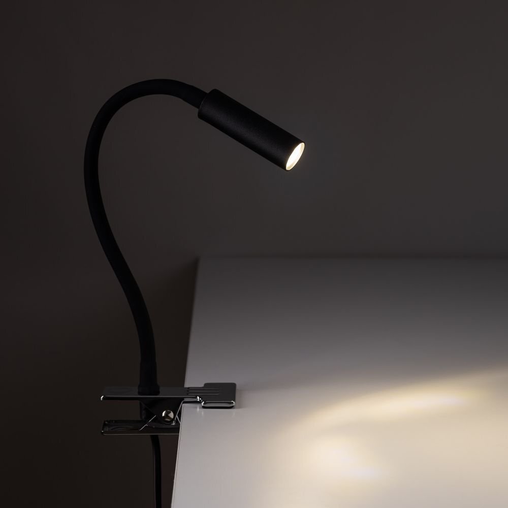 Czarna lampa biurkowa z klipsem przymocowana do krawędzi blatu, z elastycznym ramieniem i świecącym źródłem światła. Światło pada na powierzchnię blatu, tworząc jasną plamę.