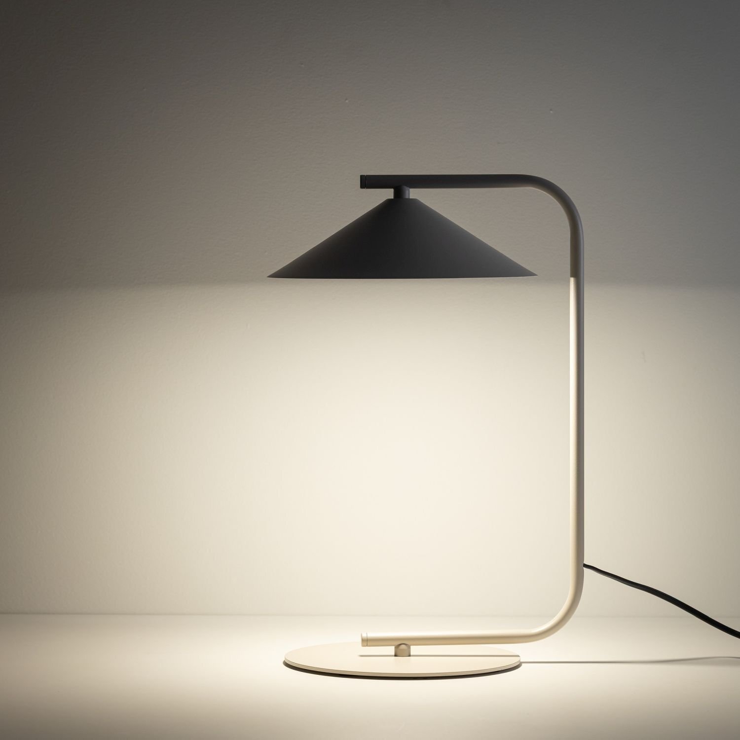 Na obrazie widoczna jest lampa biurkowa o nowoczesnym designie, z metalowym stelażem i stożkowym kloszem. Lampa emituje ciepłe światło, a jej przewód jest widoczny na powierzchni, na której stoi.
