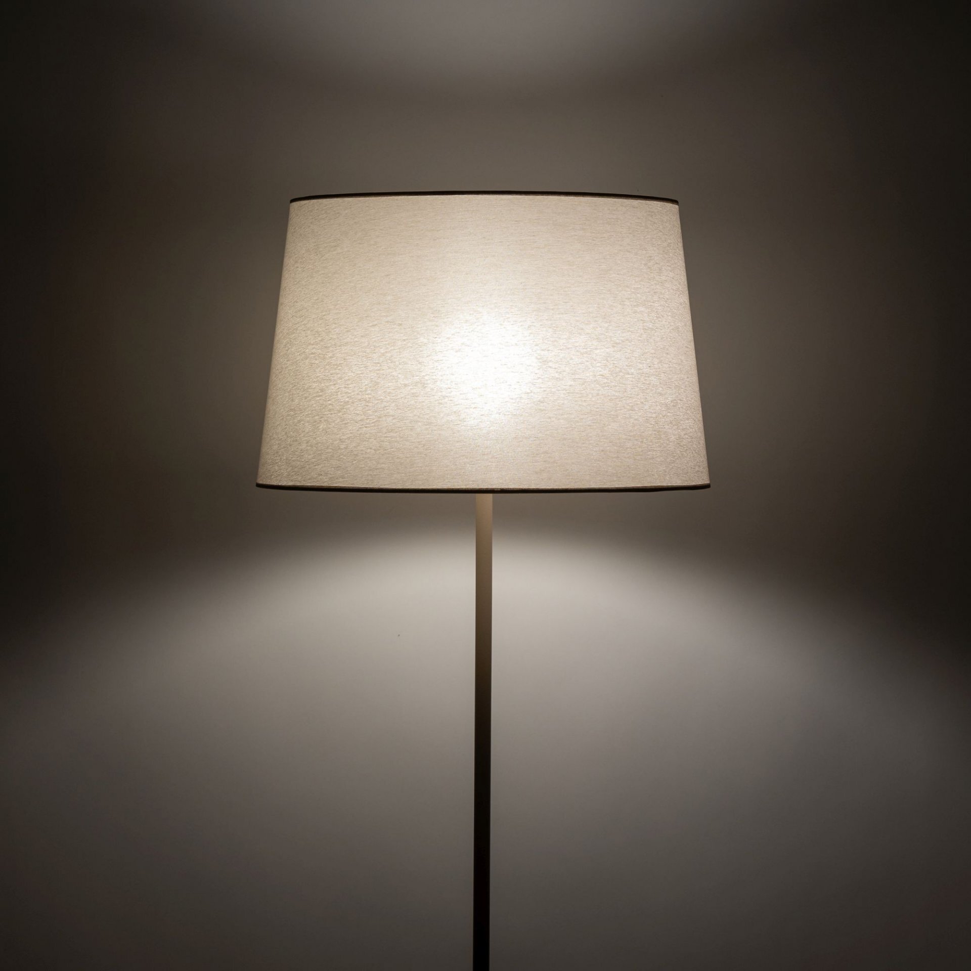 Lampa podłogowa z tkaninowym kloszem w kolorze ecru, emitująca przytulne światło. Klosz ma kształt stożka, a światło rozchodzi się równomiernie wokół niego.