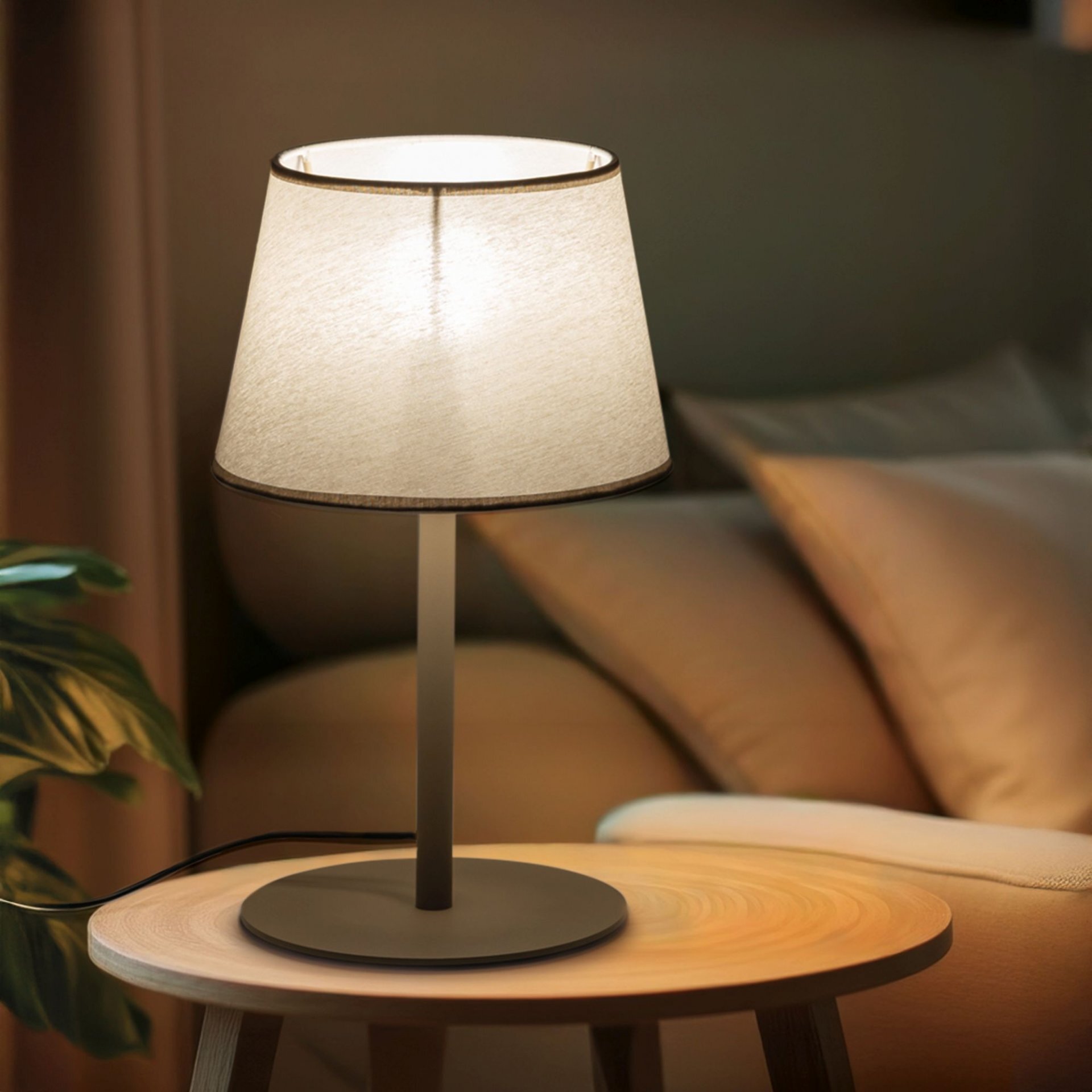 Na drewnianym stoliku stoi beżowa lampa stołowa z prostym, cylindrycznym abażurem. W tle widoczne są poduszki na sofie oraz roślina doniczkowa.