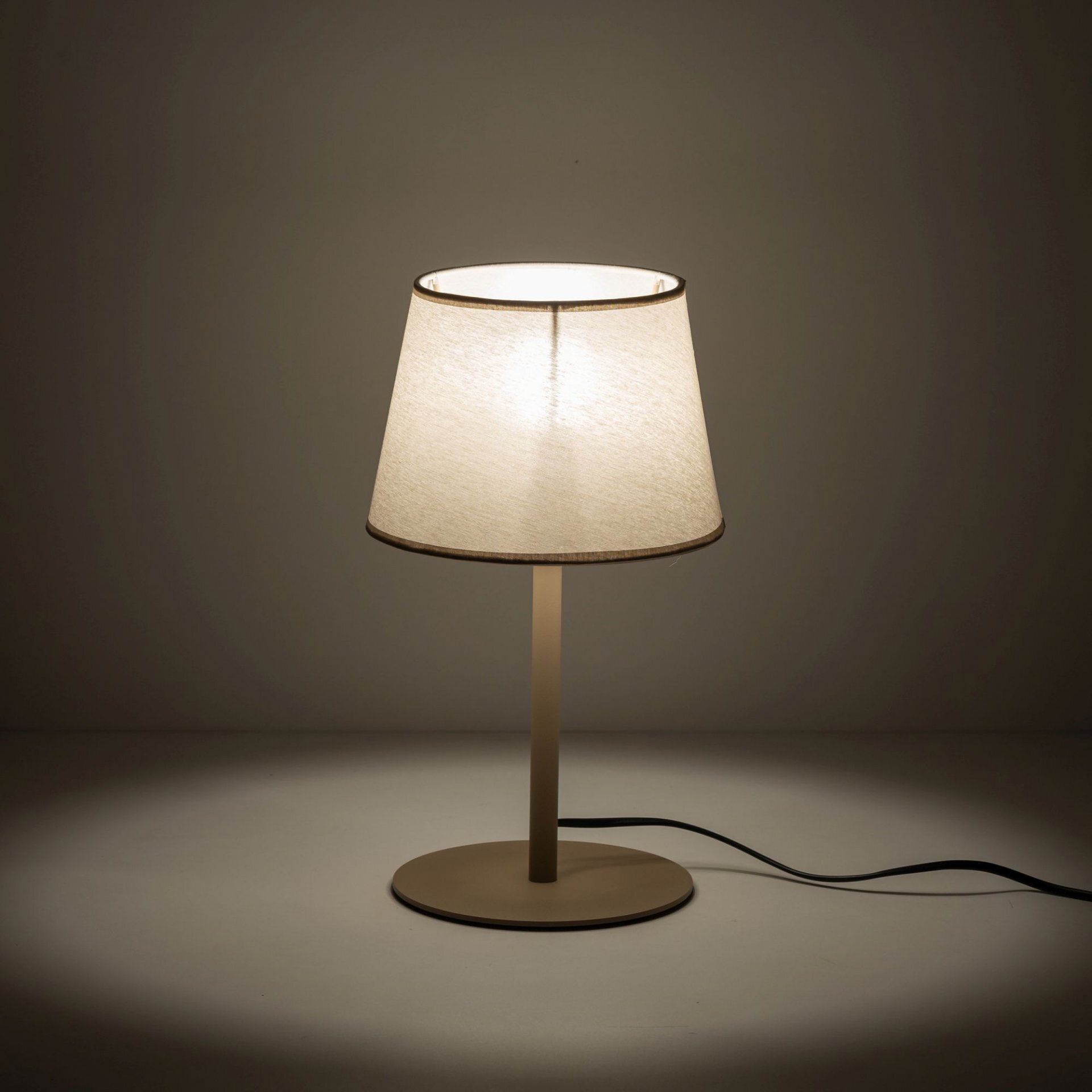 Na obrazie widoczna jest beżowa lampa stołowa z prostym abażurem, stojąca na okrągłej podstawie. Lampa jest włączona, emitując ciepłe światło, a kabel zasilający jest widoczny na powierzchni.
