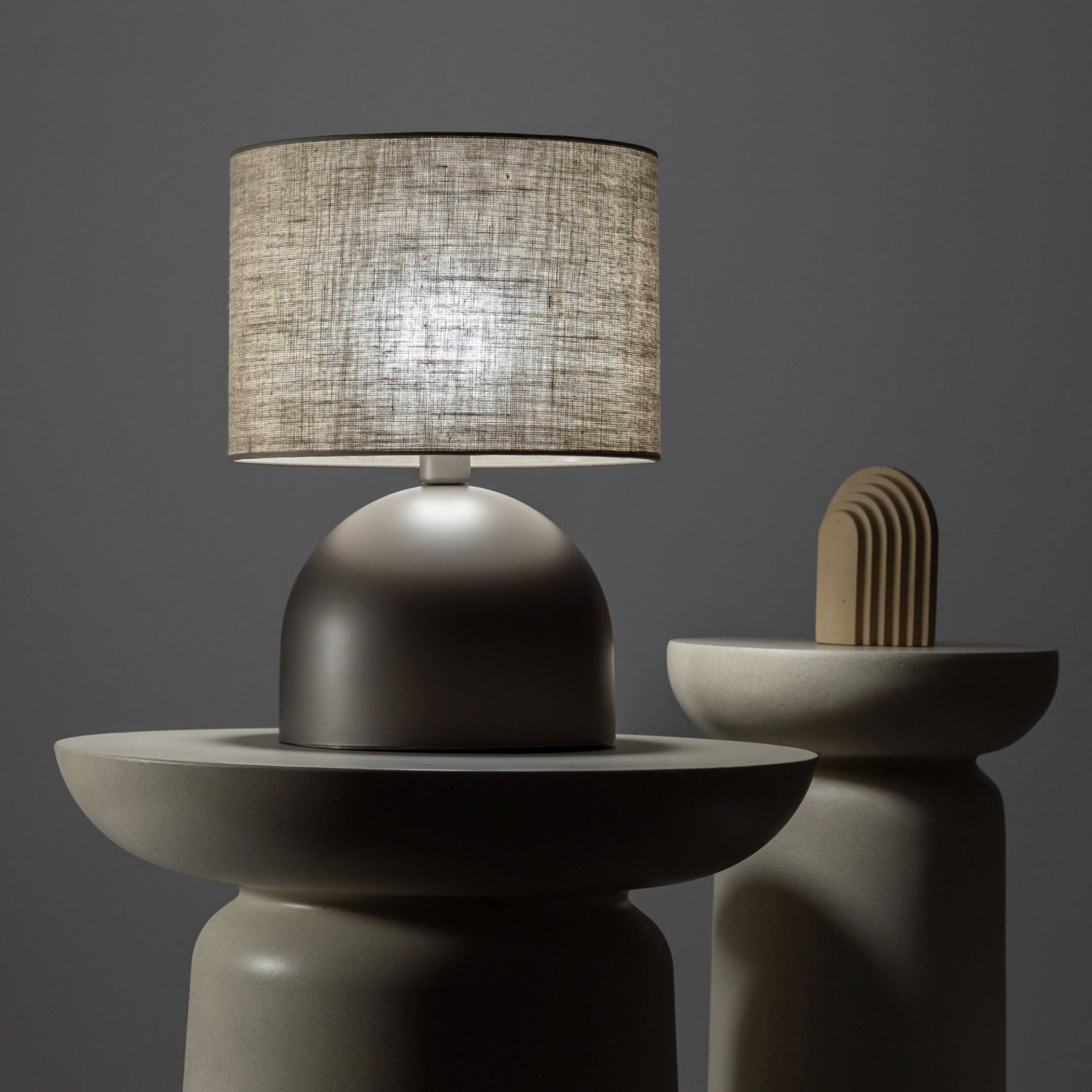 Na obrazie widoczna jest lampa stołowa z okrągłym, lnianym kloszem w kolorze brązowym, umieszczona na gładkiej, zaokrąglonej podstawie. Obok lampy znajduje się dekoracyjny element o podobnej kolorystyce.