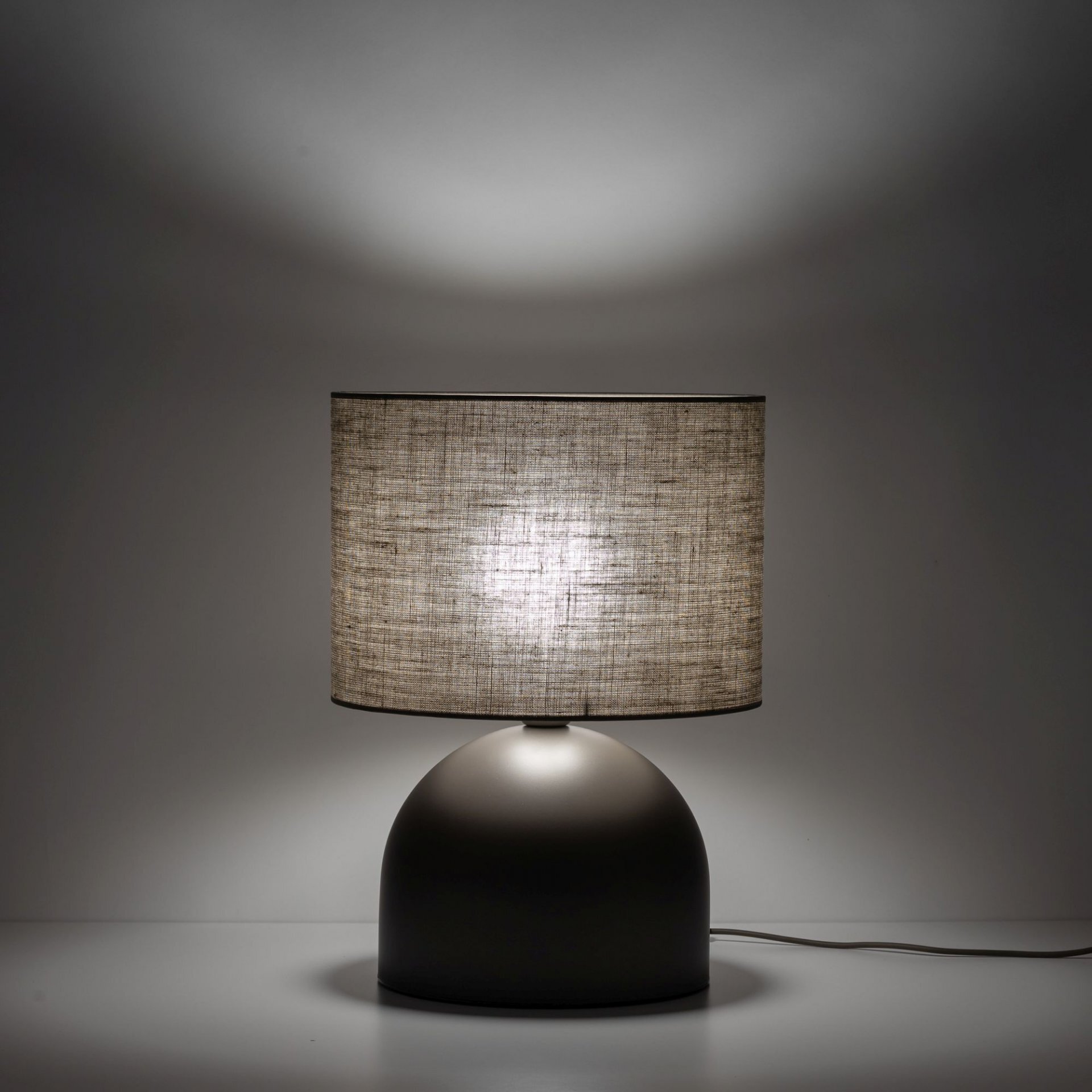 Lampa stołowa z brązowym, lnianym kloszem na półokrągłej podstawie. Światło emituje delikatny blask przez materiał klosza.