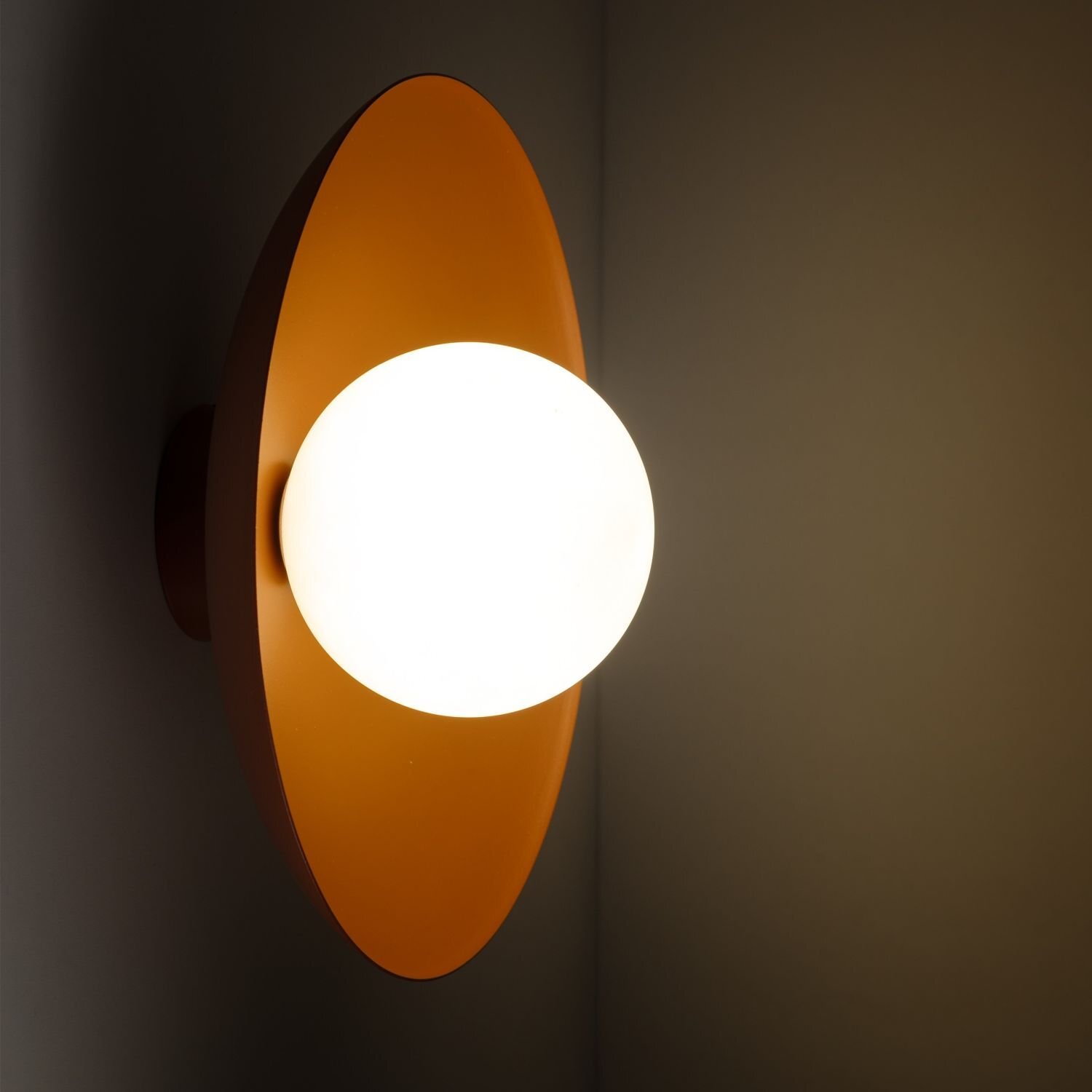 Na obrazie widoczna jest lampa z okrągłym, białym kloszem zamontowana na pomarańczowej podstawie. Światło lampy oświetla ścianę, tworząc ciepłą atmosferę.