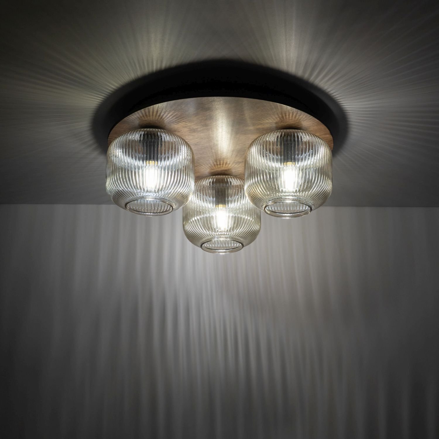 Lampa sufitowa z trzema szklanymi kloszami o żebrowanej strukturze, zamontowana na okrągłej, brązowej podstawie. Światło tworzy efektowne wzory na suficie i ścianach.
