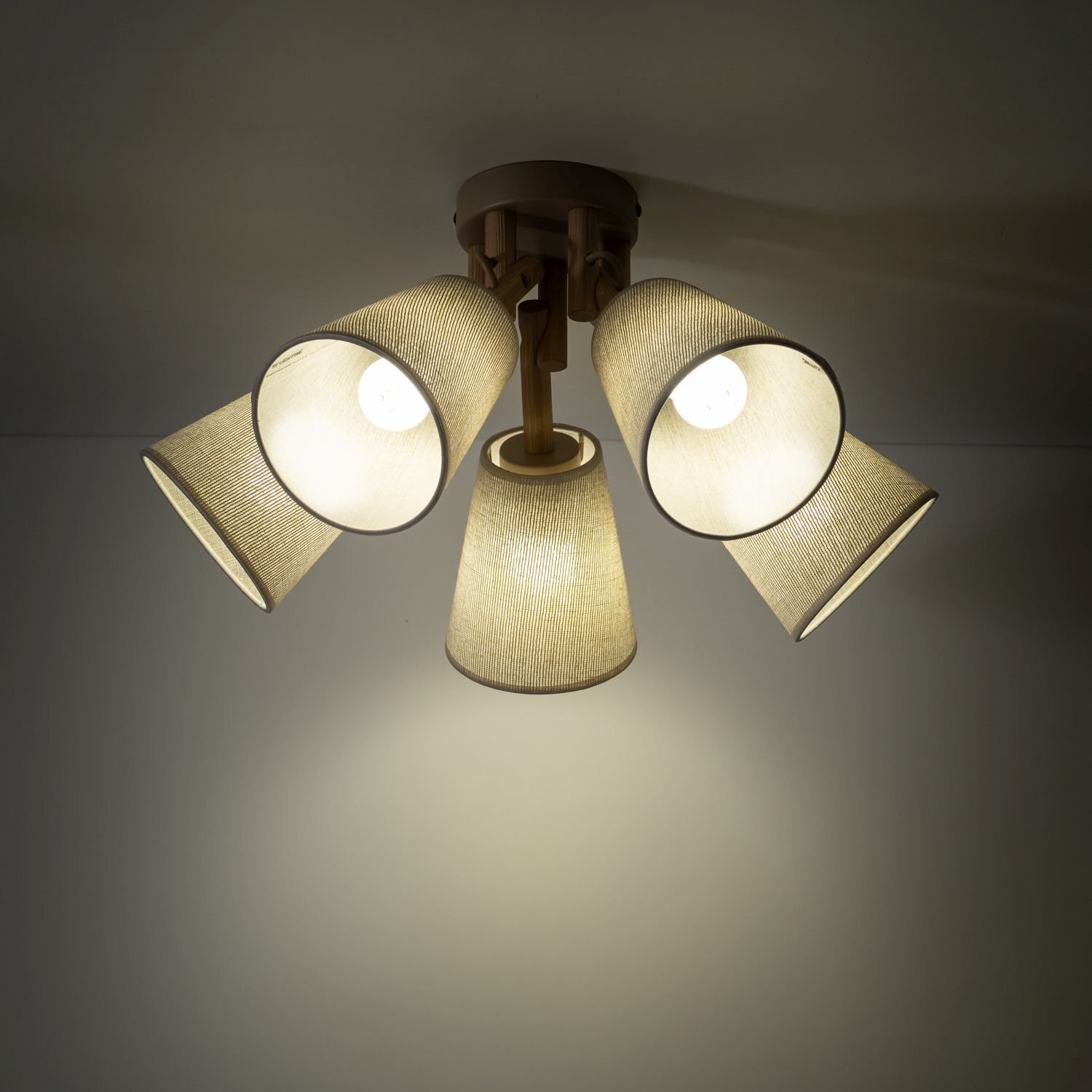 Lampa sufitowa z pięcioma kloszami w kolorze beżowym, wykonana z materiału przypominającego len, zamontowana na drewnianej podstawie. Klosze są skierowane w różnych kierunkach, a światło jest włączone.