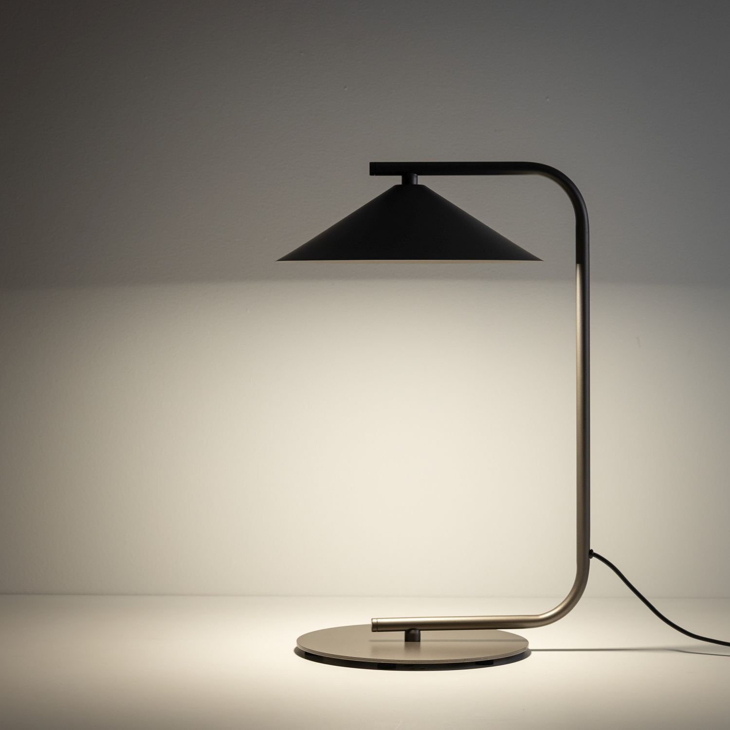 Na obrazie widoczna jest nowoczesna lampa biurkowa z metalowym stelażem i stożkowym kloszem. Lampa stoi na okrągłej podstawie, a z boku widoczny jest przewód zasilający.