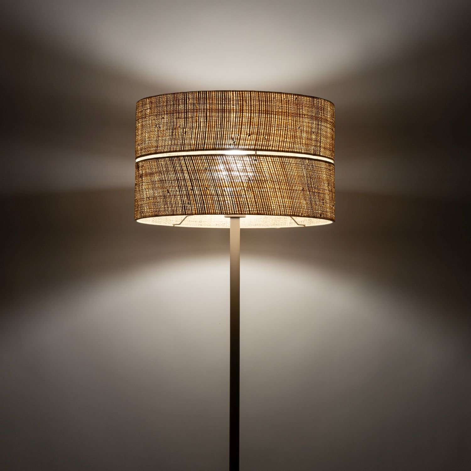 Lampa podłogowa z kloszem z rafii, emitująca ciepłe światło. Klosz ma beżowy kolor i plecioną strukturę.