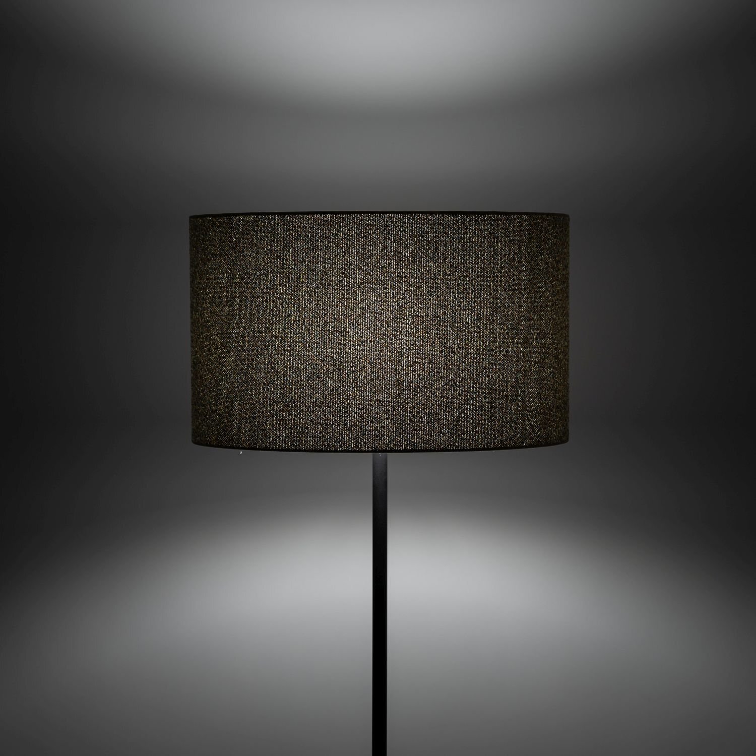 Czarna lampa podłogowa z cylindrycznym abażurem, stojąca na prostym, smukłym trzonie. Tło jest ciemne, co podkreśla elegancki wygląd lampy.