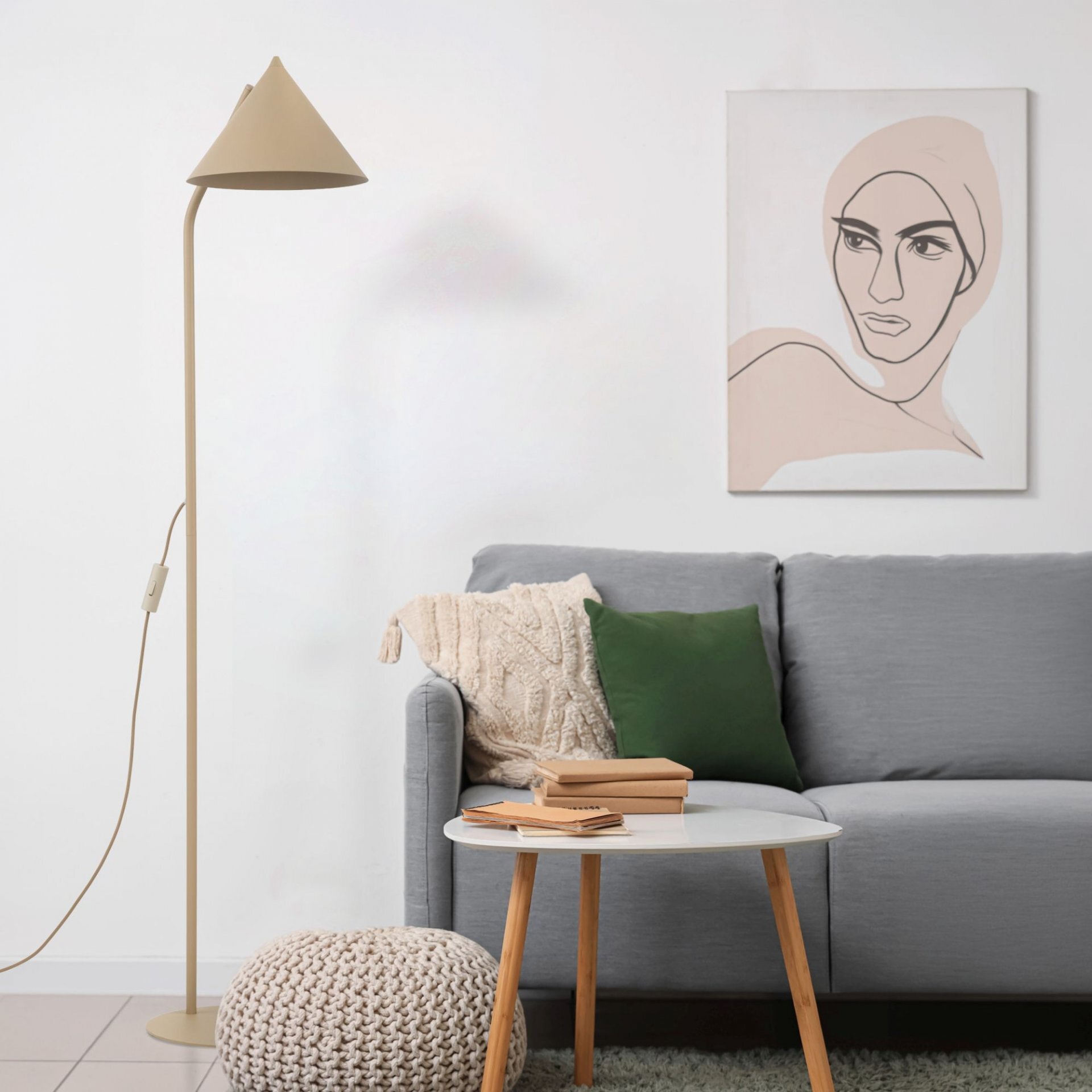 Beżowa lampa podłogowa o stożkowym kloszu stoi obok szarej kanapy z zieloną poduszką. Na ścianie wisi obraz z minimalistycznym portretem, a przed kanapą znajduje się biały stolik z książkami.
