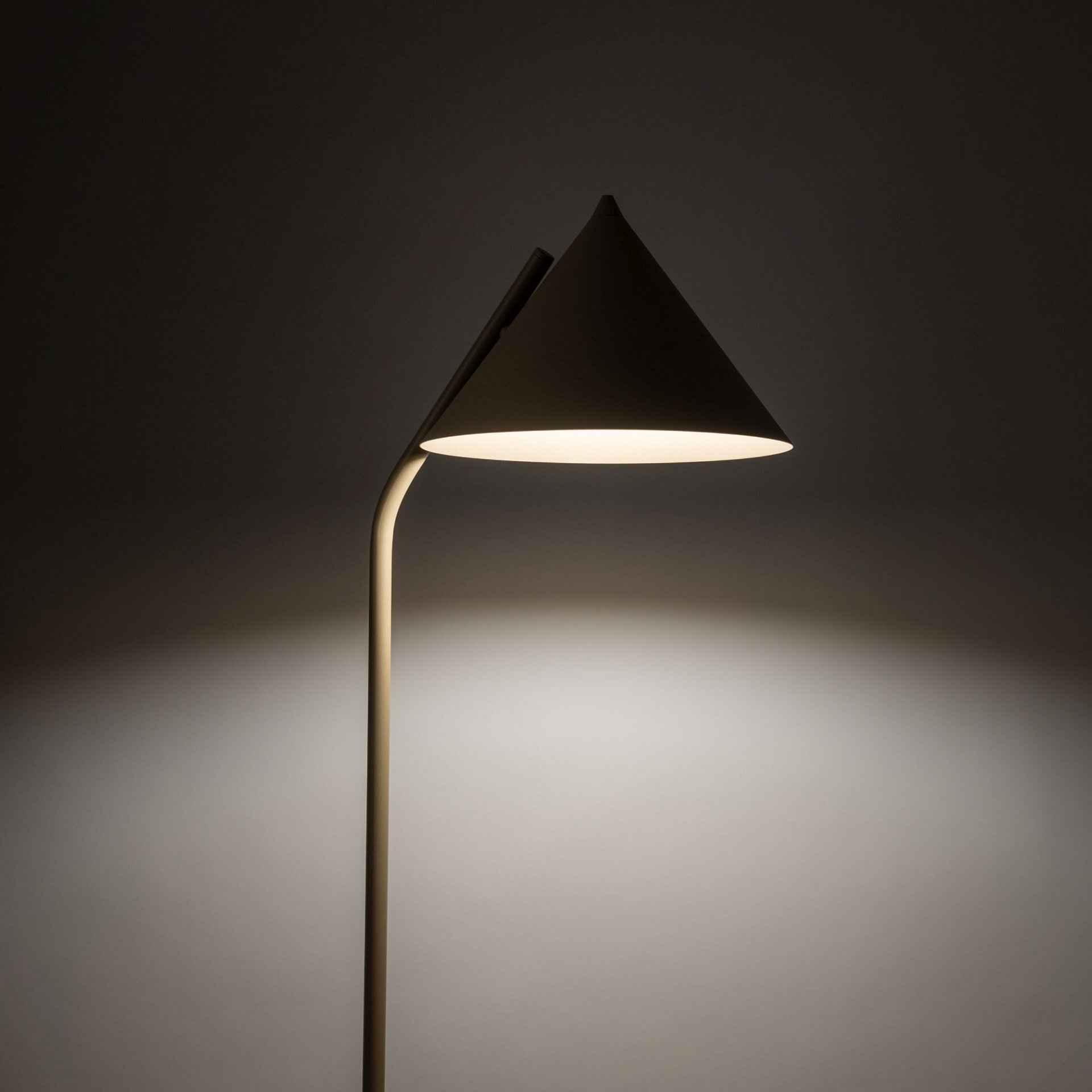 Na obrazie widoczna jest beżowa lampa podłogowa z kloszem w kształcie stożka, emitująca ciepłe światło. Lampa ma prosty, nowoczesny design i jest ustawiona na tle ciemnej ściany.