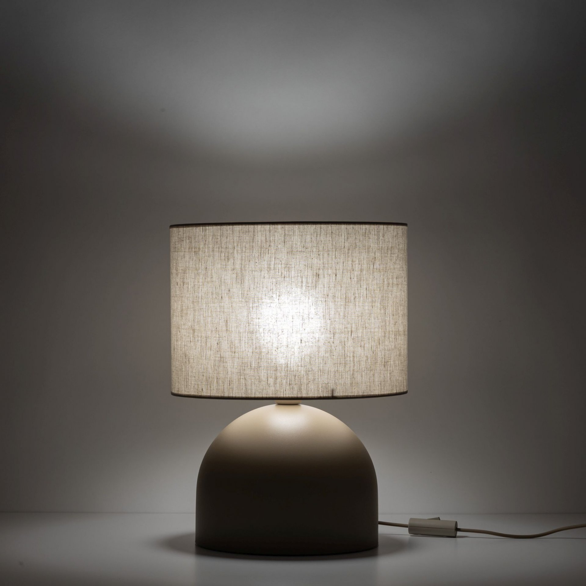 Na obrazie widoczna jest lampa stołowa z beżowym, lnianym abażurem i zaokrągloną podstawą. Lampa jest włączona, co powoduje, że abażur jest delikatnie podświetlony.
