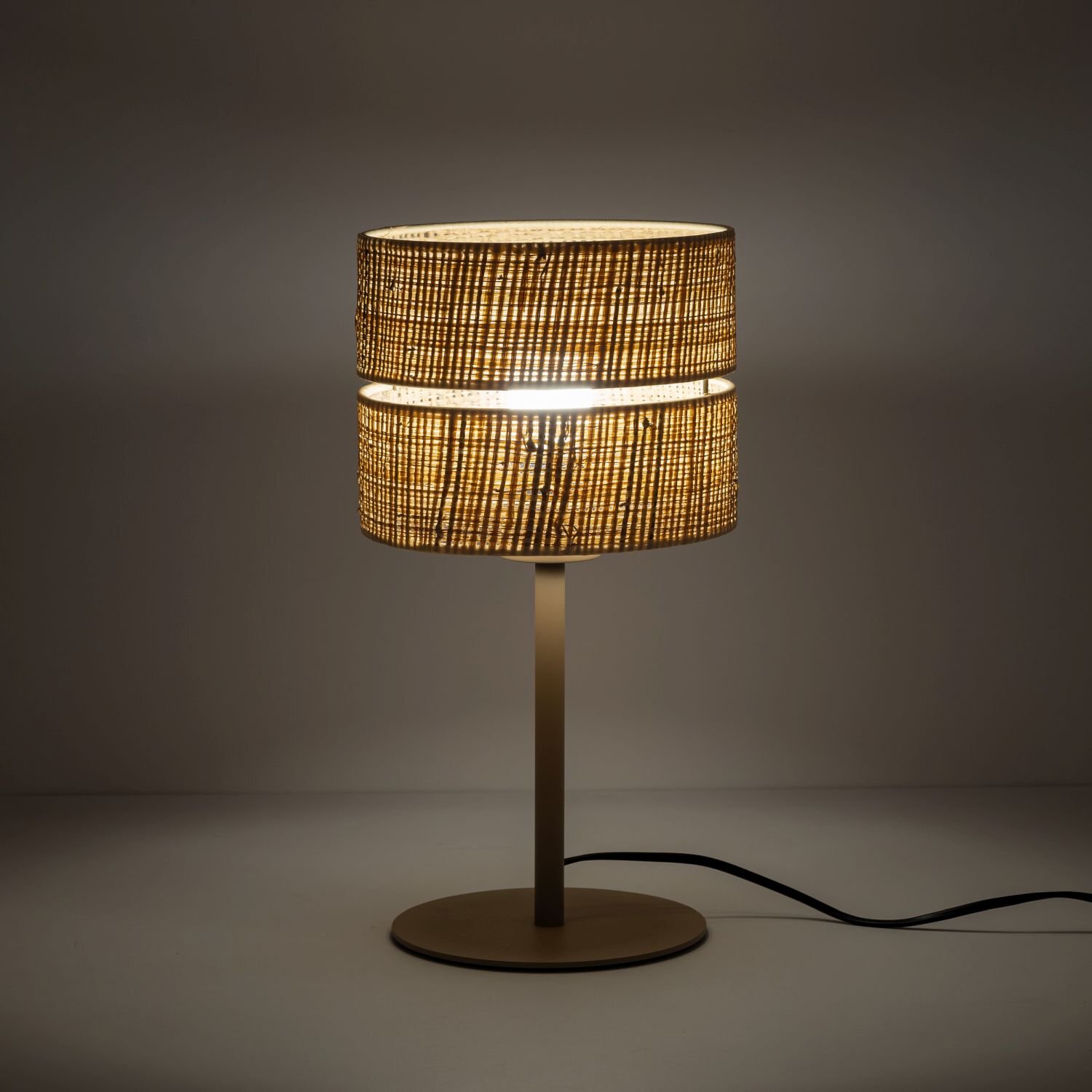 Lampa stołowa z rafiowym abażurem w kolorze beżowym, emitująca ciepłe światło. Posiada prostą, metalową podstawę i czarny przewód zasilający.