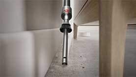 Wąska końcówka odkurzacza wciąga kurz z przestrzeni między ścianą a meblem. Podłoga jest jasna i czysta.