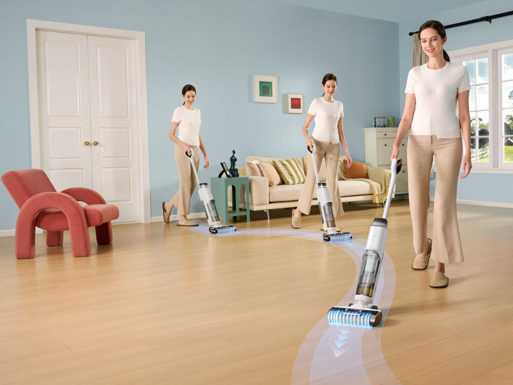 Trzy kobiety używają odkurzacza pionowego na drewnianej podłodze w jasnym salonie. W tle widać sofę, stolik kawowy i dekoracje ścienne.