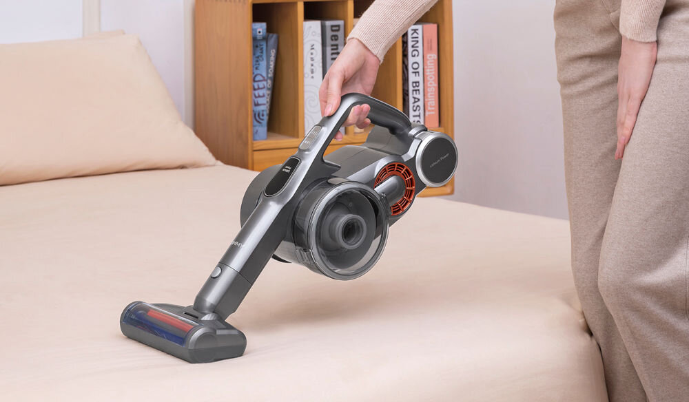 Osoba trzyma odkurzacz ręczny nad powierzchnią łóżka. W tle widoczna jest półka z książkami.