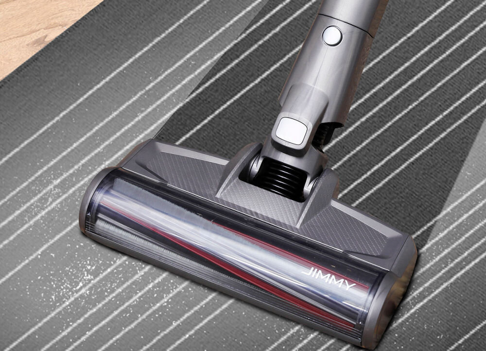 Na obrazie widoczna jest głowica odkurzacza z napisem \'JIMMY\', czyszcząca szary dywan z białymi paskami. Na dywanie znajdują się drobinki brudu.