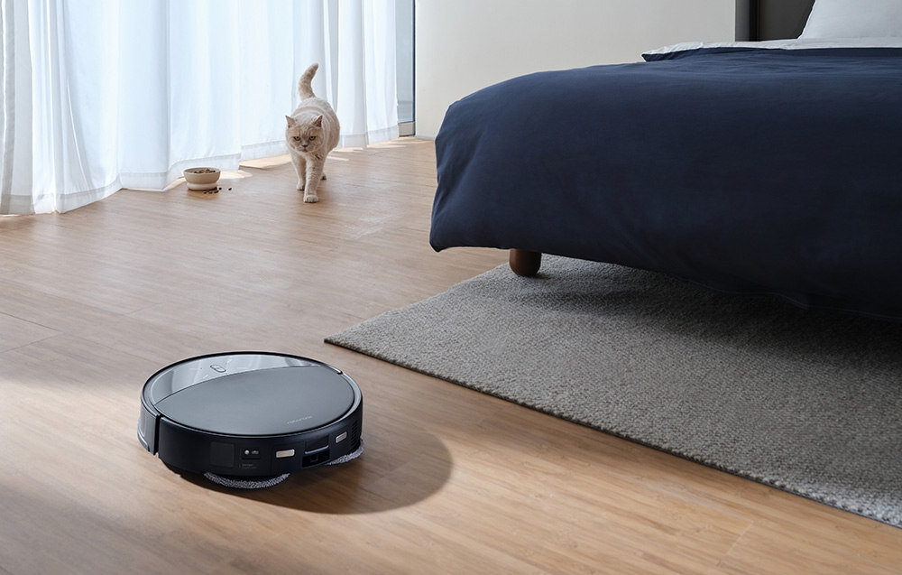 Na drewnianej podłodze w pokoju znajduje się robot sprzątający. W tle widać kota oraz łóżko z ciemną pościelą na dywanie.