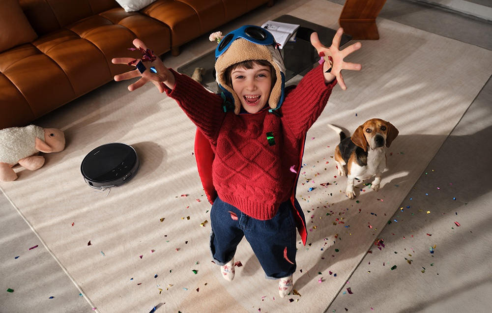 Na dywanie w salonie znajduje się robot sprzątający. Obok dziecko w czerwonym swetrze i pies, wokół nich leżą kolorowe konfetti.