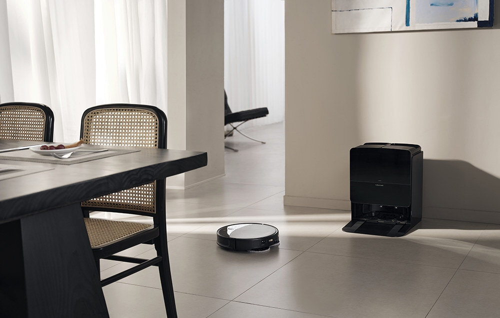 W nowoczesnym wnętrzu znajduje się robot sprzątający na podłodze oraz stacja dokująca przy ścianie. Obok stoi stół z krzesłami o plecionych oparciach.