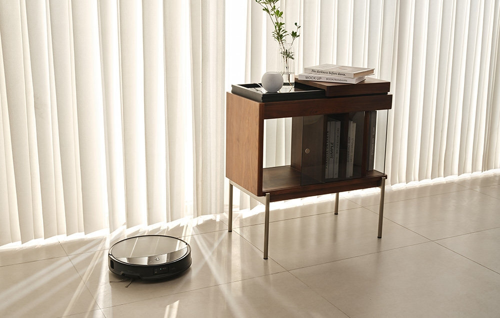 Na podłodze w jasnym pomieszczeniu znajduje się robot sprzątający. Obok stoi drewniana szafka z książkami i wazonem z gałązką.