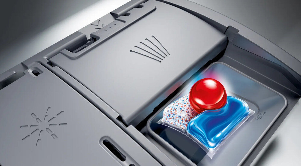 Wnętrze zmywarki z otwartym pojemnikiem na detergent, w którym znajduje się kapsułka do zmywania z czerwonym i niebieskim żelem. Obok pojemnika widoczne są wytłoczone symbole wskazujące na funkcje urządzenia.