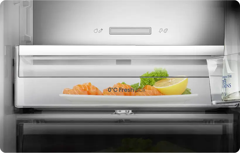 Wnętrze lodówki z widoczną szufladą oznaczoną jako \'0°C Fresh BOX\', w której znajduje się talerz z kawałkami łososia, cytryną i sałatą. Obok szuflady stoi butelka z napisem \'FROM THE MOUNTAINS\'.