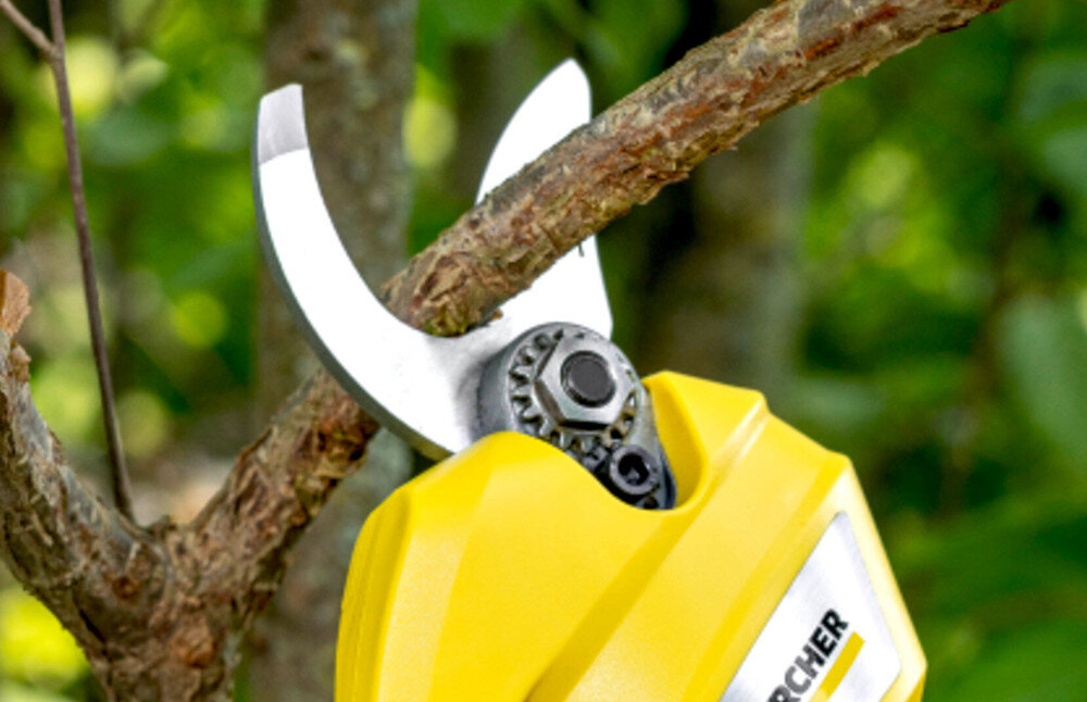 Na obrazie widoczny jest żółty sekator tnący gałąź drzewa. Na urządzeniu znajduje się logo \'KARCHER\'.
