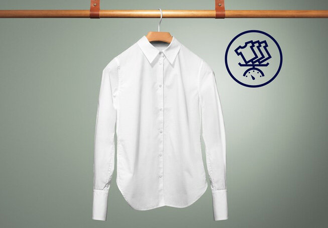 Biała koszula wisi na drewnianym wieszaku na tle zielonej ściany. Obok koszuli znajduje się niebieska ikona przedstawiająca trzy koszule nad zegarem.