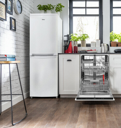 W kuchni znajduje się biała lodówka z logo \'Amica\' oraz otwarta zmywarka w zabudowie. Na blacie stoją rośliny doniczkowe i różne akcesoria kuchenne.