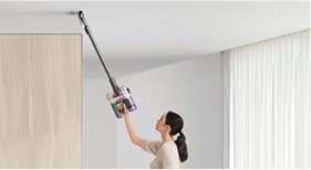 Osoba używa odkurzacza pionowego do czyszczenia sufitu w jasnym pomieszczeniu z zasłonami i drewnianą ścianą.