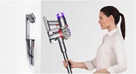 Osoba trzyma odkurzacz pionowy z fioletowym elementem na górze, obok widać uchwyt montażowy na ścianie. Tło jest jasne i jednolite.