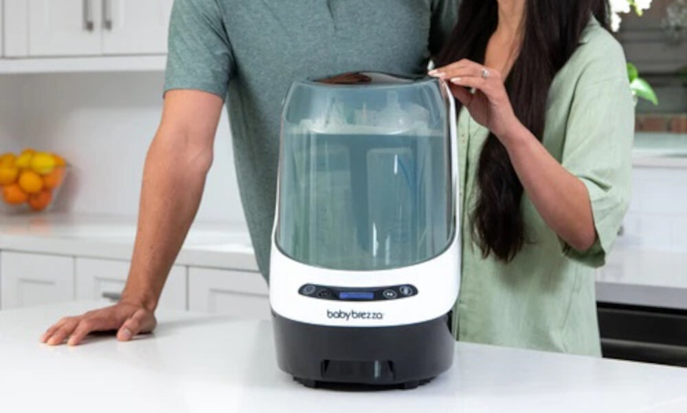 Na blacie kuchennym stoi sterylizator Baby Brezza Bottle Washer Pro w kolorze czarnym. Obok urządzenia znajdują się dwie osoby, których twarze nie są widoczne.