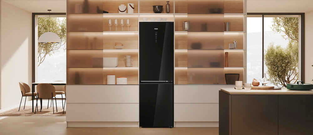 Czarna lodówka z błyszczącym wykończeniem stoi w nowoczesnej kuchni z drewnianymi półkami i dużymi oknami. Na lodówce widoczne jest logo \'Haier\'.